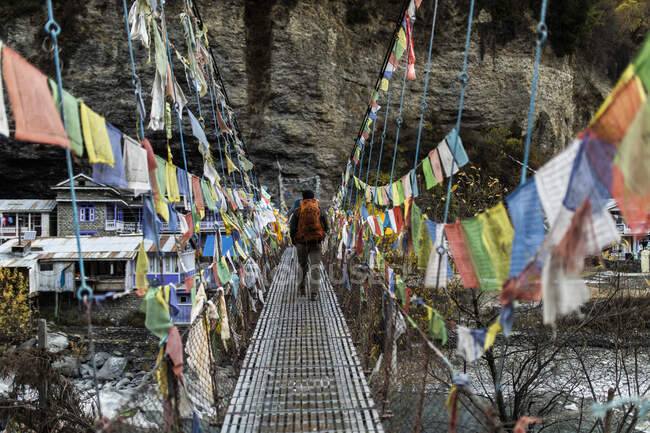 Visão traseira do viajante irreconhecível com mochila andando ao longo de passarela de suspensão metálica decorada com bandeiras de oração coloridas no Himalaia, no Nepal — Fotografia de Stock