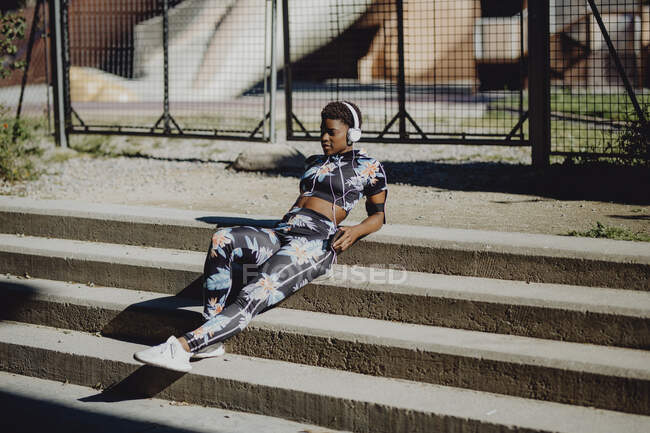 Jeune femme afro-américaine en vêtements de sport colorés écouter de la musique dans les écouteurs tout en étant assis sur des escaliers en pierre dans le parc — Photo de stock