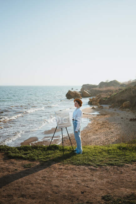Вид збоку молодої жінки в стильному одязі і береті, що стоїть на трав'яному узбережжі біля піску і океану в сонячний день, малюючи картину пензлем на мольберті — стокове фото