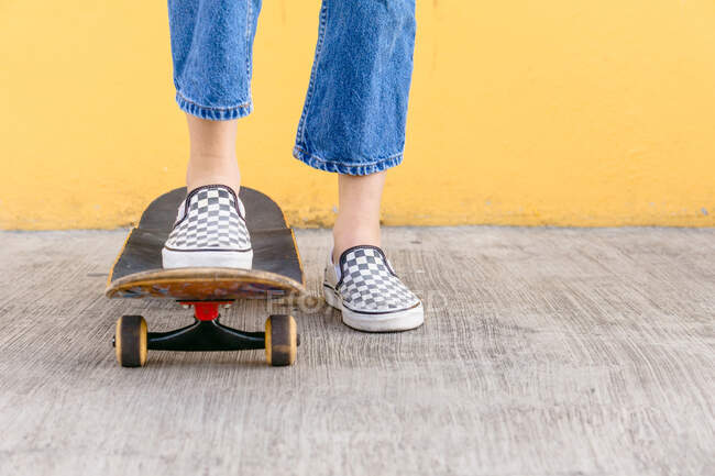 Ausgeschnittene unkenntliche Skaterin mit Skateboard steht tagsüber auf Gehweg mit bunter gelber Wand auf dem Hintergrund — Stockfoto