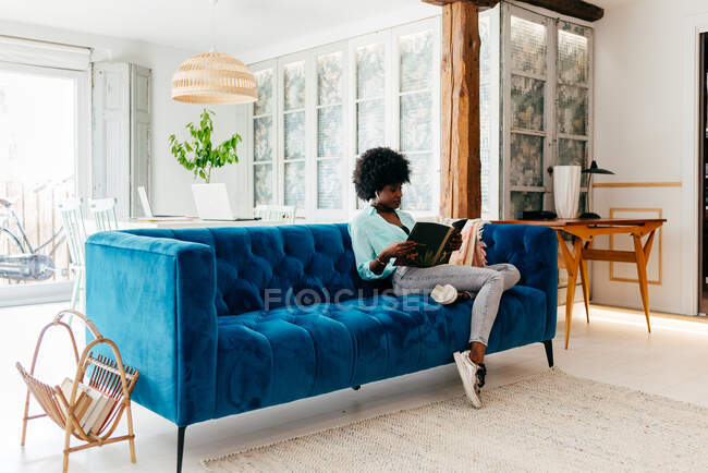 Junge schwarze Frau in lässigem Outfit mit Kopfhörern sitzt auf gemütlichem blauem Sofa zu Hause und liest Magazin, während sie die Freizeit zu Hause genießt — Stockfoto
