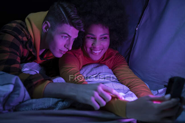 Счастливые мультирасовые мужчина и женщина, улыбающиеся отдыхая и просматривая мобильный телефон в палатке ночью — стоковое фото