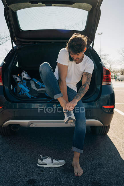 Tattooed male athlete putting on footwear while talking on cellphone against car with open trunk in city — Stock Photo