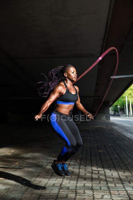 Vista lateral de la hermosa mujer afroamericana en ropa deportiva sosteniendo la cuerda de salto y mirando a la cámara mientras está parado en el pavimento en la calle de la ciudad - foto de stock