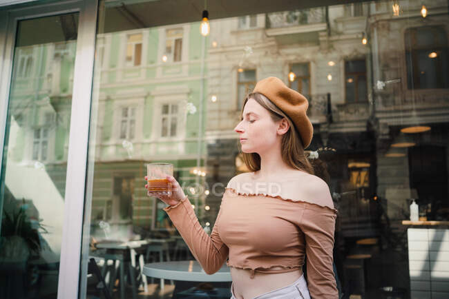 Stylish serene female in trendy beret standing with glass of coffee near window of cafe in street — Stock Photo