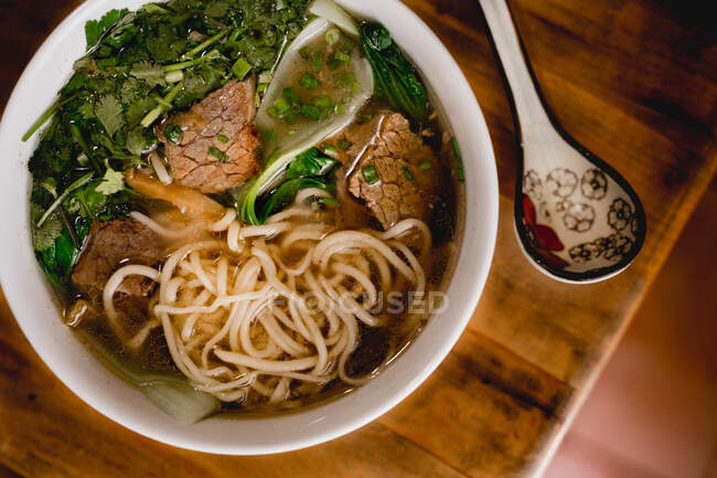 From above big white ceramic bowl of beef ramen soup and colorful spoon placed on wooden table — Stock Photo