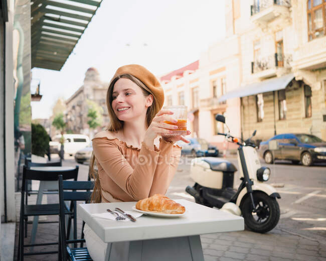 Französin in Baskenmütze sitzt am Tisch im Café mit aromatischem Glas Kaffee und frisch gebackenem Croissant — Stockfoto