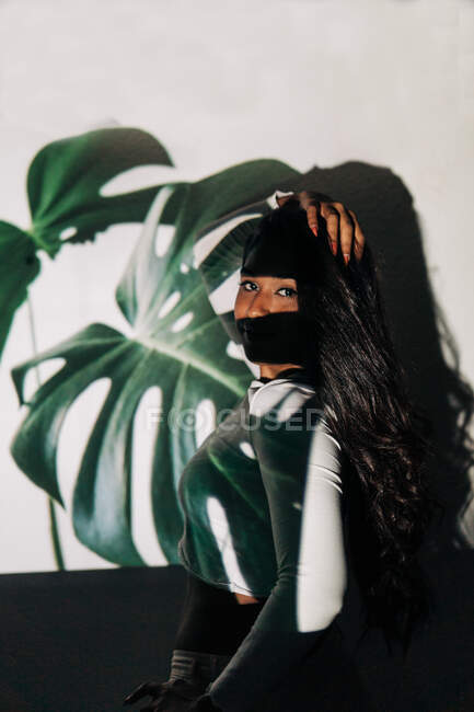 Jovem hispânica fêmea tocando o cabelo e olhando para a câmera enquanto estava de pé contra a parede com projeção de folha de planta monstera — Fotografia de Stock