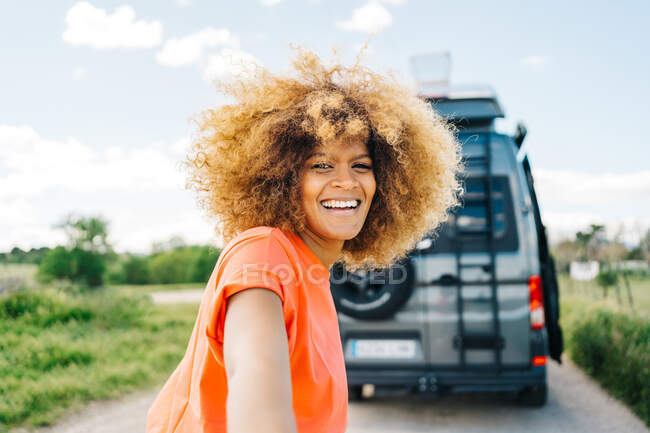 Feliz mujer afroamericana con longboard sonriendo y mirando a la cámara mientras conduce amigo de la cosecha a la camioneta durante el viaje por carretera - foto de stock
