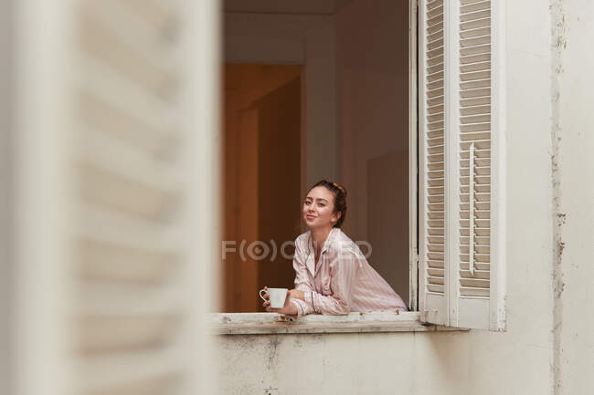 Tranquil hembra en pijama inclinada en la ventana con taza de café de la mañana y mirando a la cámara - foto de stock