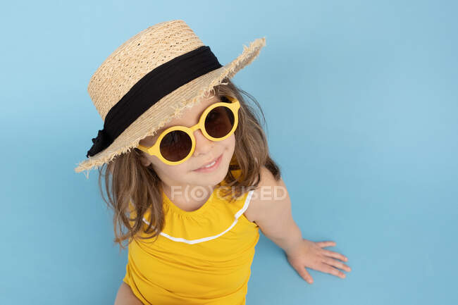 Высокий угол милой счастливой маленькой девочки в жёлтом купальнике и соломенной шляпе со стильными солнечными очками, сидящей на синем фоне и смотрящей в камеру — стоковое фото