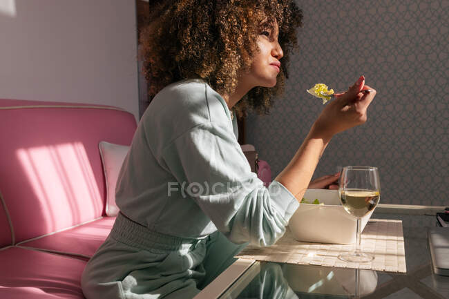Vue latérale de la femme afro-américaine mangeant de la salade assis à table avec un verre de vin et ayant un délicieux déjeuner à la maison — Photo de stock
