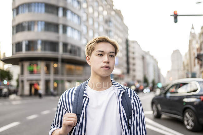 Moda modelo masculino asiático com cabelo loiro e mochila de pé na rua da cidade e olhando para longe — Fotografia de Stock