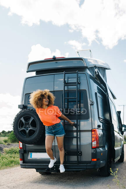 Optimistische Afroamerikanerin mit lockigem Haar lächelt und schaut weg, während sie an einem Sommertag auf dem Land die Leiter auf dem Rücken des Wohnmobils greift — Stockfoto