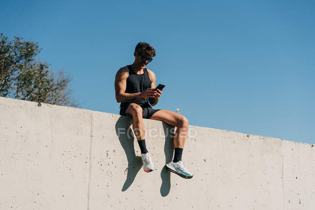 Ajuste atleta masculino en ropa deportiva que navega por el teléfono celular mientras descansa en la valla después del entrenamiento - foto de stock