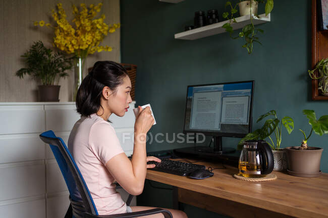 Vista lateral de la freelancer asiática leyendo documentos en el monitor de la computadora mientras está sentada en la mesa y bebiendo té durante el trabajo remoto - foto de stock