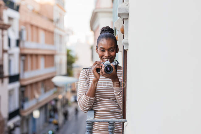 Содержание этнической женщины в одежде с полосатым орнаментом с профессиональным фотоустройством, смотрящей в камеру на балконе днем — стоковое фото