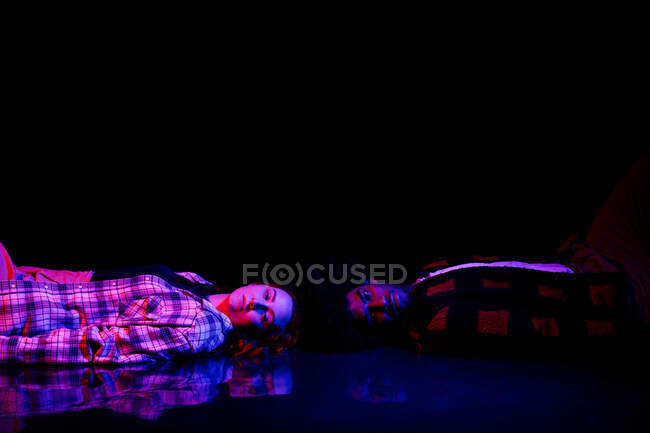 Seitenansicht von aufgebrachten jungen Männern und Frauen in karierten Hemden, die in einem dunklen Raum mit Neonbeleuchtung auf dem Boden liegen und in die Kamera schauen — Stockfoto
