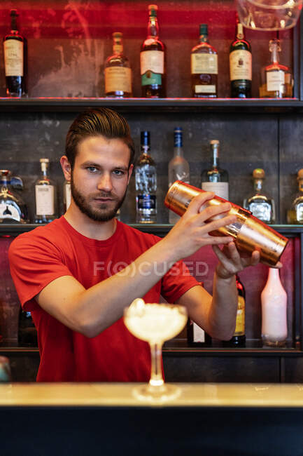 Орієнтований бармен готує алкогольний коктейль і змішуючи інгредієнти в шейкері, стоячи за лічильником в барі і дивлячись на камеру — стокове фото