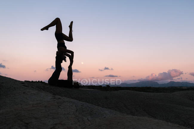 Vista laterale della donna flessibile che si equilibra a testa in giù mentre pratica acroyoga con il partner maschile contro il cielo al tramonto in montagna — Foto stock
