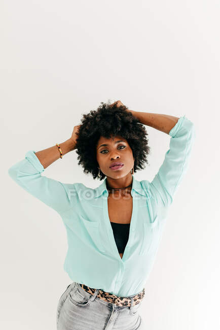 Художня молода афроамериканка в модному одязі торкається волосся афро, дивлячись на біле тло. — стокове фото