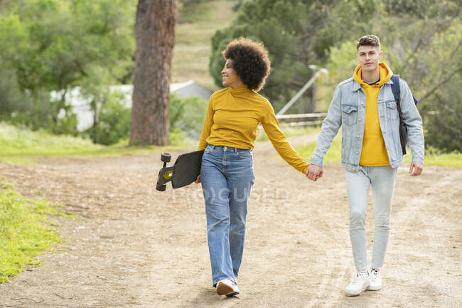 Full body diverse young man and woman with longboard holding hands and walking on countryside road on summer weekend day — Stock Photo