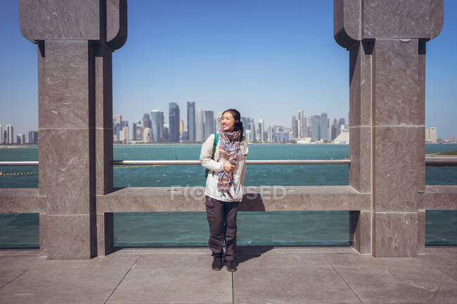 Передній вид радісної азіатської жінки-туристки посміхається в камері, стоячи один біля аркової огорожі на набережній Музею ісламського мистецтва проти Західної затоки і прекрасного краєвиду центру в Доха. — стокове фото