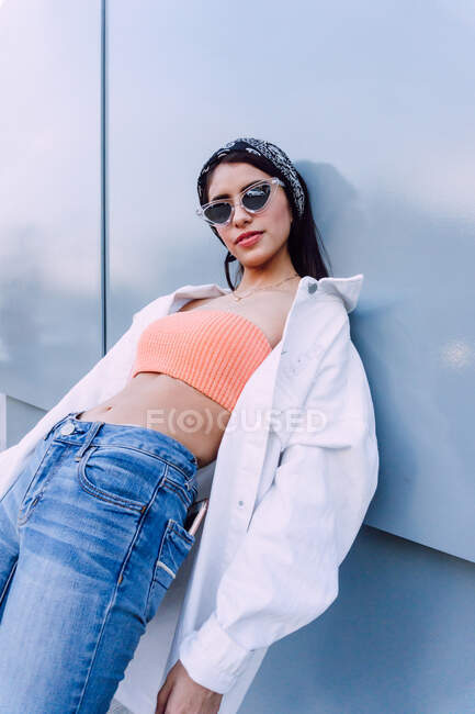 Mulher confiante vestindo jeans com camisa superior e branca com óculos de sol elegantes em pé perto da parede — Fotografia de Stock