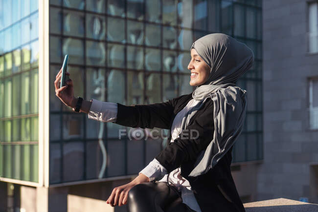 Visão lateral da mulher muçulmana positiva no hijab tomando auto-retrato no smartphone na cidade no dia ensolarado — Fotografia de Stock