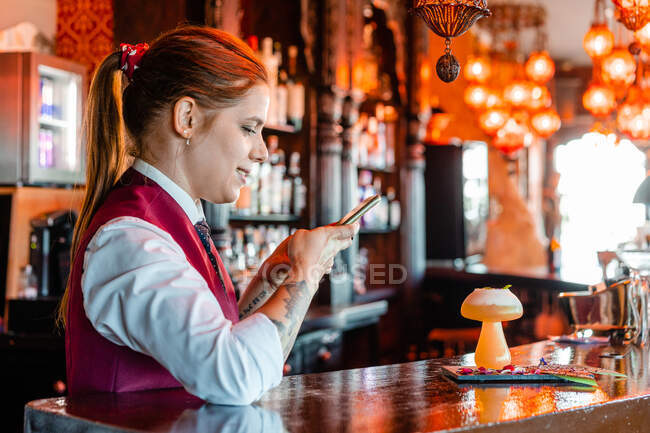Вид сбоку на веселую барменшу, фотографирующую кислый коктейль в стакане, подаваемый на стойке в пабе — стоковое фото