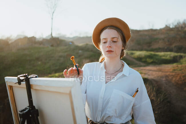 Молода жінка в стильному одязі і береті стоїть на трав'яному узбережжі біля піску і океану в сонячний день, малюючи картину пензлем на мольберті — стокове фото