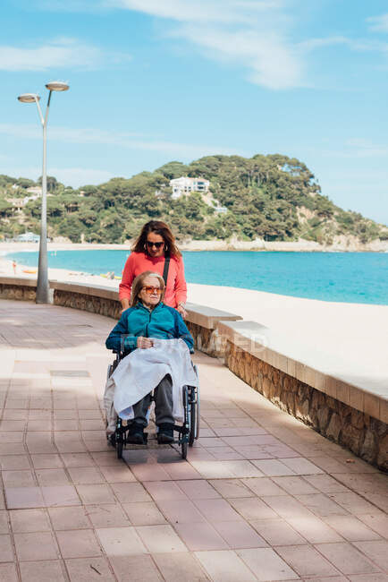 Erwachsene Tochter läuft im Sommer mit älterer Mutter im Rollstuhl entlang der Strandpromenade am Meer — Stockfoto