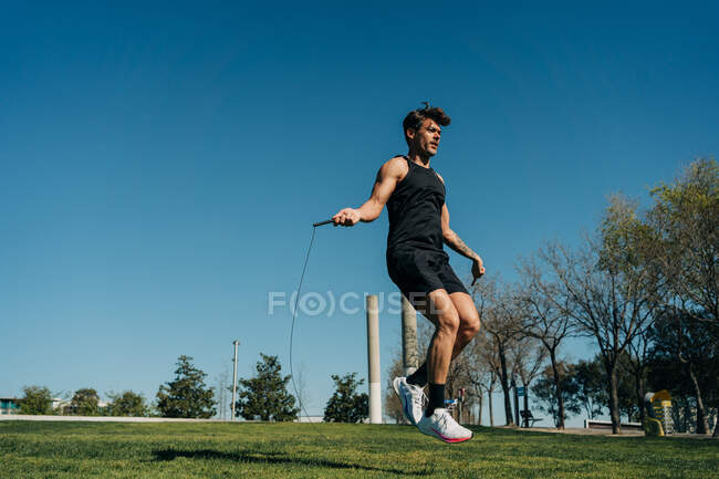 Atleta masculino em sportswear pulando com corda e olhando para longe na passarela durante o treinamento cardio no parque — Fotografia de Stock