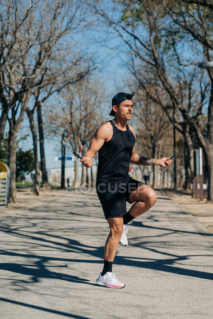 Atleta di sesso maschile in abbigliamento sportivo che salta con corda da salto e distoglie lo sguardo sulla passerella durante l'allenamento cardio nel parco — Foto stock