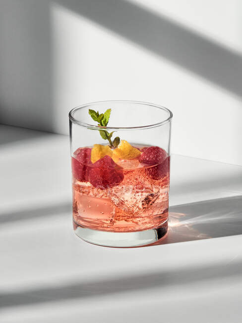 Glass with cocktail with ice cubes and raspberries with mint sprig placed on white surface — Stock Photo