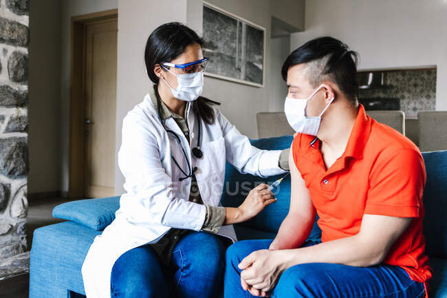 Female medic with syringe making injection of vaccine for Latin teenage boy with Down syndrome at home during coronavirus — Stock Photo
