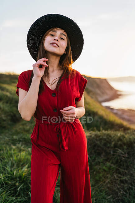 Jolie jeune femme en robe de soleil rouge et chapeau debout sur une prairie herbeuse verdoyante dans une campagne ensoleillée — Photo de stock