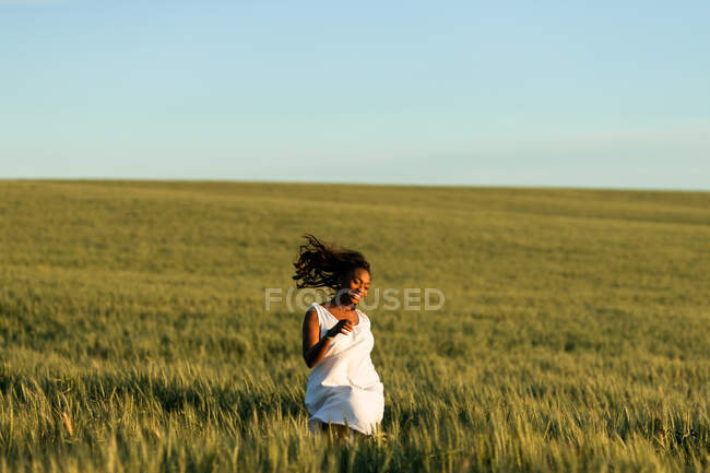 Sonriente joven dama negra en vestido de verano blanco paseando por el campo de trigo verde mientras mira hacia otro lado durante el día bajo el cielo azul - foto de stock