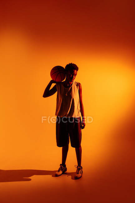Mujer negra con traje de baloncesto en el estudio usando geles de color y luces del proyector sobre fondo naranja - foto de stock
