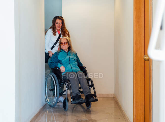 Mujer adulta satisfecha empujando silla de ruedas con madre mayor y caminando por el pasillo en el centro de rehabilitación - foto de stock