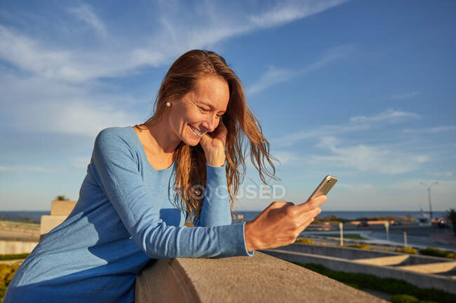 Улыбающаяся взрослая леди просматривает телефон, опираясь на забор возле океана на городской улице в солнечный день — стоковое фото