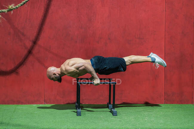 Боковой вид босоногого мужчины, делающего упражнения на брусьях о красную стену во время тренировки в тренажерном зале — стоковое фото