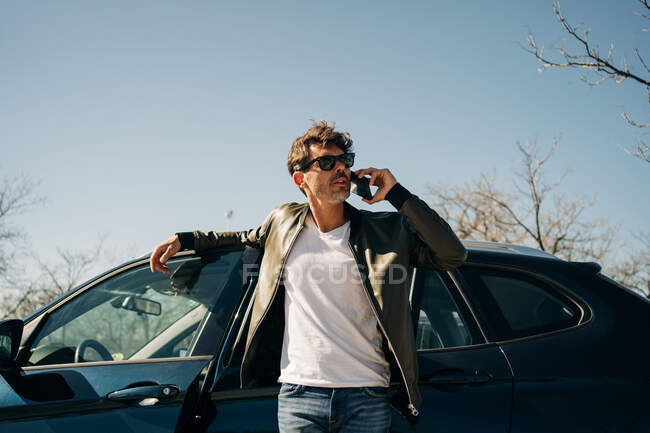 Чоловік в сонцезахисних окулярах говорить на мобільному телефоні, стоячи біля автомобіля в сонячний день — стокове фото