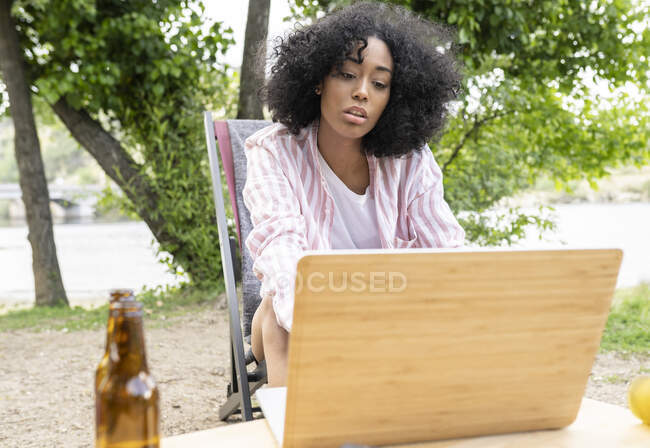 Jeune femme hispanique avec les cheveux bouclés assis près de la table avec ordinateur portable le week-end à la campagne — Photo de stock