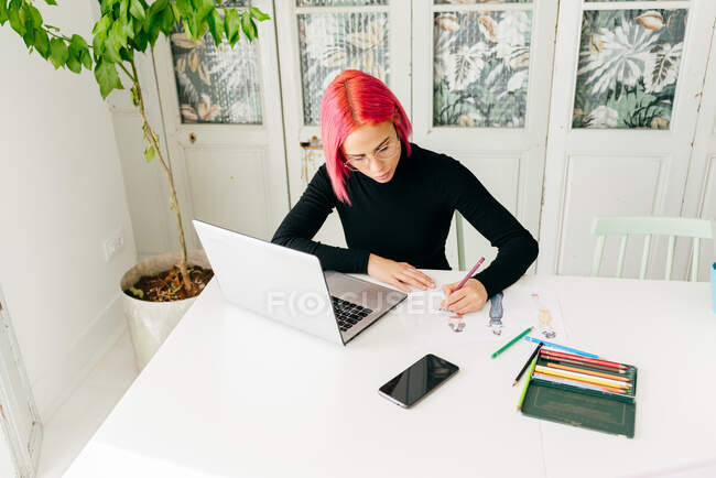 Von oben eine nachdenkliche freiberufliche Designerin in lässigem Outfit und Brille, die am Tisch mit Laptop arbeitet und Modeskizzen mit Bleistiften zeichnet — Stockfoto