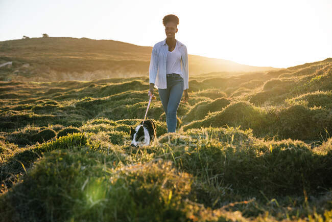 Full body of happy ethnic woman with Border Collie dog walking together on trail among grassy hills in sunny spring evening — Stock Photo