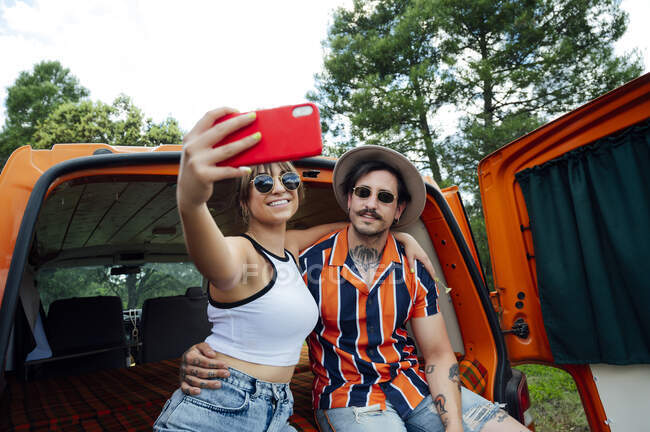 Casal encantado de viajantes sentados em van e tomando auto-tiro no smartphone enquanto abraça e desfruta de aventura de verão — Fotografia de Stock