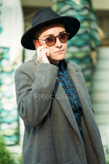 Side view of androgynous person in hat and modern sunglasses talking on cellphone while looking at camera standing on the street in daylight — Stock Photo