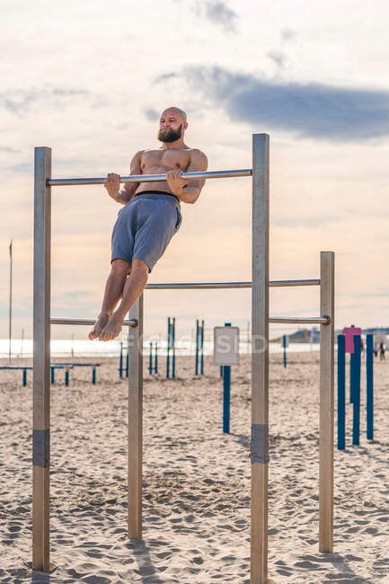 Shirtless bearded man doing muscle up on chin up bar while training on sunny beach — Stock Photo