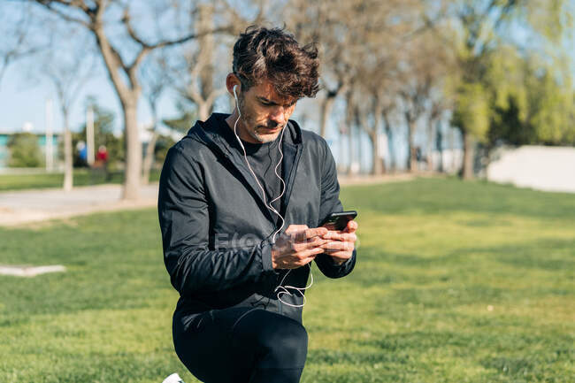 Bearded male athlete in sportswear and earphones text messaging on cellphone while squatting on meadow in town — Stock Photo